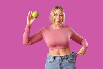 Deurstickers Pretty young woman in loose jeans with apple on pink background. Weight loss concept © Pixel-Shot