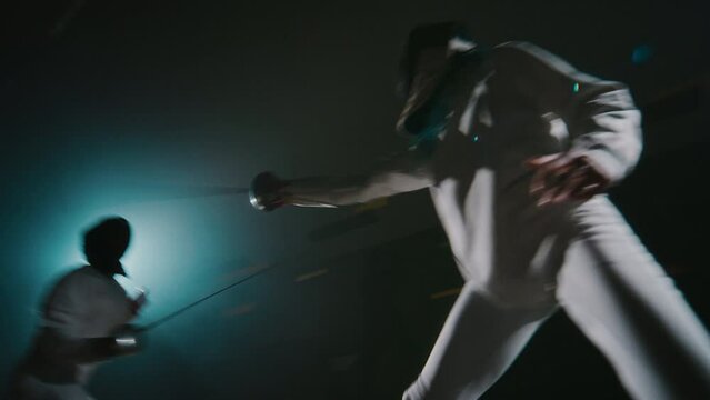 Side handheld footage of two foil fencers dodging and riposting during tough fencing match on rapiers in dark hall