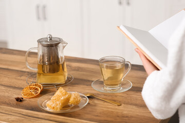 Composition with teapot, cup of tea and young woman reading book in kitchen