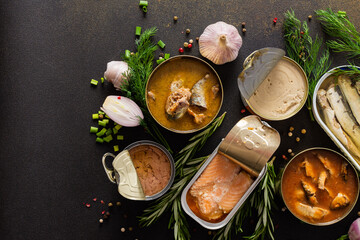 Different open tin cans with canned fish among spices and herbs on a brown background, canned salmon and mackerel, sprat and sardine, tuna and herring and fish pate, top view