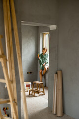 Young woman paints walls while making repairment of a new apartment. Having a break and sitting on a window sill. Creative process of home renovation and repair concept - 763311861