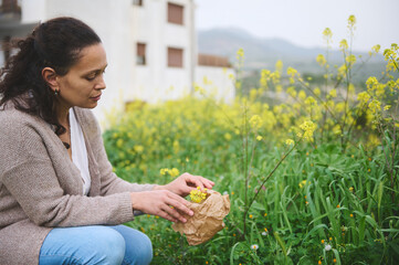Confident young woman gathering herbs and plants for medical use, collecting flowers in a paper bag...