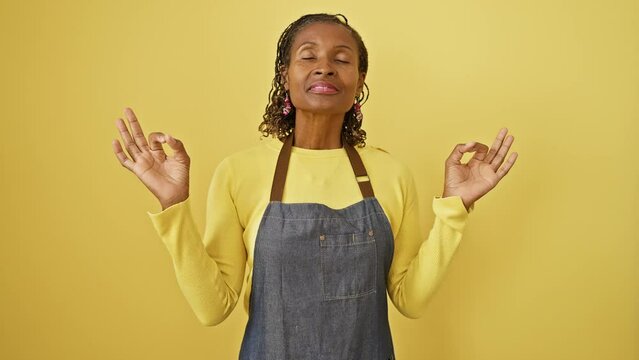 Radiant middle-aged african american woman, apron-clad, engaging in peaceful yoga meditation while standing on a vibrant, isolated yellow backdrop