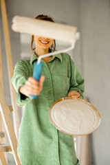 Portrait of a young joyful and cute woman standing with paint roller during repairing process of a house. Concept of happy leisure time while renovating interior - 763313006