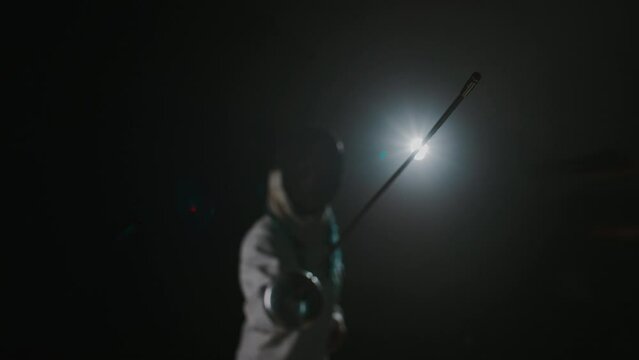 Medium footage of foil fencer pointing fencing rapier to camera while standing against black background with light
