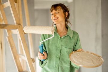 Portrait of a young joyful and cute woman standing with paint roller during repairing process of a house. Concept of happy leisure time while renovating interior