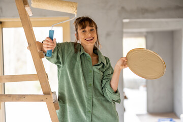 Portrait of a young joyful and cute woman standing with paint roller during repairing process of a house. Concept of happy leisure time while renovating interior - obrazy, fototapety, plakaty