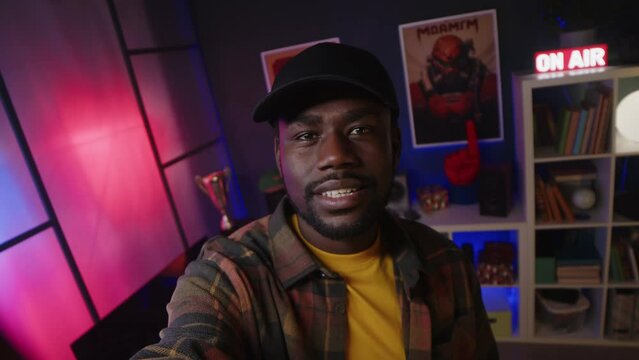 Handheld POV of young African American guy wearing cap and checkered shirt recording himself on camera for video blog while spending night time in neon light gamer room
