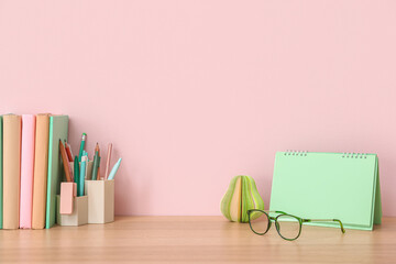 Books, stationery supplies, flip calendar and glasses on wooden table near pink wall