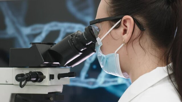 A microbiologist in glasses looks into a microscope, analyzing a sample. Female scientist working with high-tech equipment.