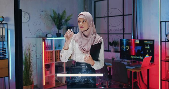 Woman office worker in hijab which standing near glass wall and making important notes on it in evening office
