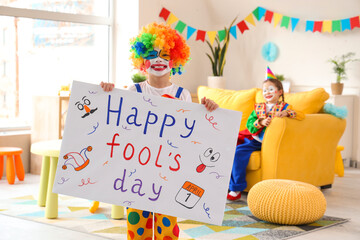 Funny little boy in clown costume with poster at home. April Fools' Day celebration