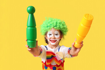 Funny little girl in clown costume with juggling clubs on yellow background. April Fools' Day...