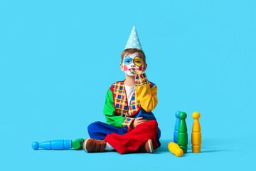 Funny little boy in clown costume with party whistle and juggling clubs on blue background. April...