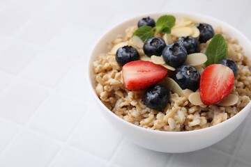 Tasty oatmeal with strawberries, blueberries and almond petals in bowl on white tiled table, closeup. Space for text