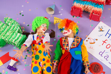 Funny little children in clown costumes with party decor on lilac background, top view. April Fools' Day celebration