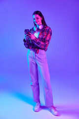 Full-length image of smiling young woman in checkered shirt using mobile phone for work and personal needs against purple background in neon. Concept of youth, lifestyle, casual fashion, emotions