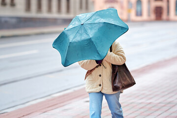 Woman strugle with gusts of wind. Woman holding an umbrella navigating the windy city streets, strong wind blowing. Female with turquoise umbrella. Person with umbrella hiding from strong stormy wind