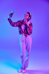 Full-length image of smiling young woman in checkered shirt talking on video call via mobile phone against purple studio background in neon. Concept of youth, lifestyle, casual fashion, human emotions
