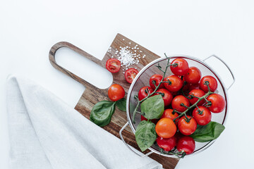 red cherry tomatoes on branches in a colander with basil leaves, on a wooden board with coarse salt...