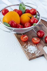 red cherry tomatoes on branches and yellow tomatoes in a colander with basil leaves, on a wooden board with coarse salt and tomato halves on a white background.