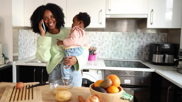 Multi-tasking Mom Balancing Childcare and Work Calls - A vibrant young mother holding her child while engaging in a phone conversation in the kitchen.