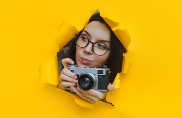 A young paparazzi girl in glasses with a rare SLR camera looks out from her hiding place and carefully looks at what is happening. Yellow paper hole. Tabloid press.Looking for a story for stock photos - obrazy, fototapety, plakaty
