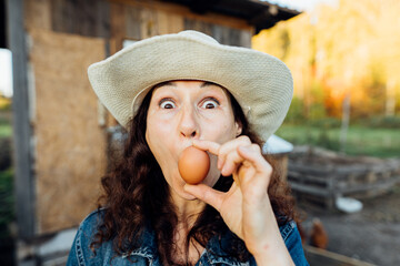 Funny Woman Farmer in denim with a fresh egg, making a funny face. A woman in hat exhibits sheer happiness with a fresh egg in hand, standing on her farm Sustainable lifestyle, organic farmer