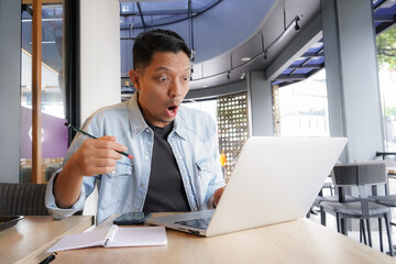 Asian man shocked face, wow, surprised with blue shirt using laptop and mobile phone in coffee...