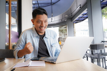 Asian man happy and enthusiastic with blue shirt using laptop and mobile phone in coffee shop,...