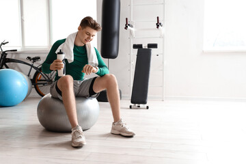 Fototapeta na wymiar Sporty young man with water bottle looking at smartwatch on fitball in gym