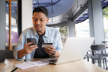 Asian man using laptop and mobile phone for shopping online with credit card in coffee shop online freelance business