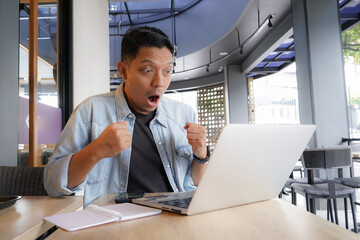 Asian man shocked face, wow, surprised with blue shirt using laptop and mobile phone in coffee shop, shopping online