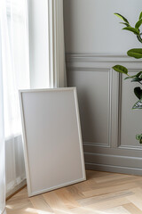 Obraz premium A white frame leaning against the wall in an empty room, with light grey walls and a parquet floor, a green plant near the window, a closeup view of a picture on the ground, from an angled view