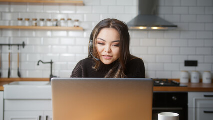 Asian woman wear wireless headset having video conference call on laptop computer talk by webcam in online chat in kitchen at home. Customer support service, distance online teaching concept	
