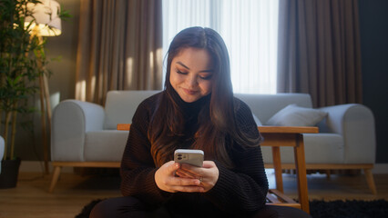 Young asian girl sitting on floor typing text message by smartphone. Woman relaxing in living room...