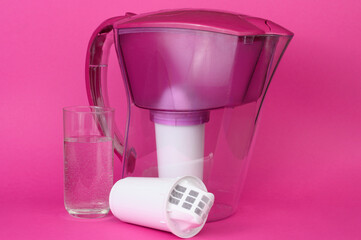 Water purification jug with spare filter and a glass of purified drinking water on pink background....