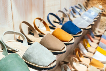 A shelf displays a variety of handmade leather sandals in various pastel colors, which show the Menorcan craftsmanship and traditional summer footwear style of the island.