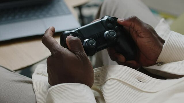 Unrecognizable Black hands of gamer pressing buttons on black controller while playing fps video game on laptop at home