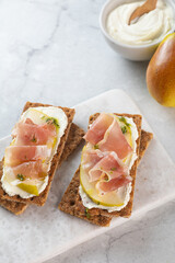 Two wholewheat crispbread with ricotta cheese, prosciutto, sliced pear and thyme on light grey marble cutting board. Healthy snack concept. Italian cuisine