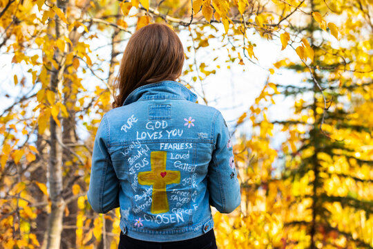 Close-up view from behind of a woman wearing a jean jacket with hand-written Christian references during a fall outing in a city park; Edmonton, Alberta, Canada