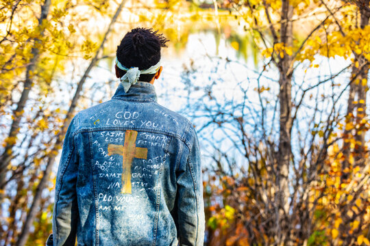 Close-up view from behind of a man wearing a jean jacket with hand-written Christian references during a fall outing in a city park, standing by a lake; Edmonton, Alberta, Canada