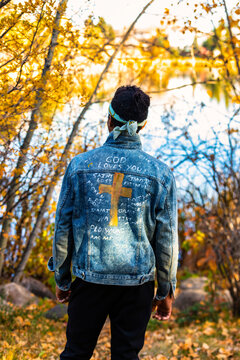 Close-up view from behind of a man wearing a jean jacket with hand-written Christian references during a fall outing in a city park, standing by a lake; Edmonton, Alberta, Canada