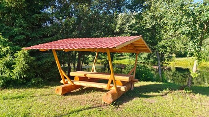 A wooden gazebo with benches, a table and a metal roof stands on a grassy lawn surrounded by spruce and apple trees. A small lake is located nearby. Sunny summer weather and blue sky