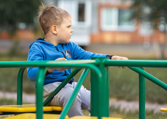 An adorable Caucasian blond boy is sitting on a green merry-go-round swing in his backyard.
