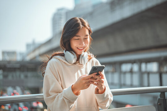 Beautiful young happy asian woman with headphones looking at mobile phone while standing in the city