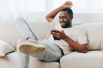 Happy African American man sitting on a black sofa, typing a message on his mobile phone The modern...