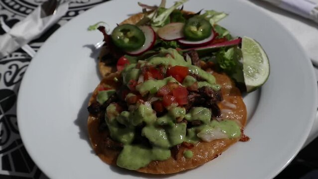 carne asada vampiro tostada and fish taco on a plate at a mexican restaurant (green avocado sauce, lime, white plate) garnished with radishes, jalapeno peppers, pico de gallo, tomatos onions cilantro