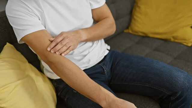 Young hispanic man scratching arm indoors, feeling discomfort while sitting in a living room at home.