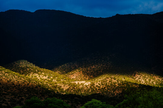A shaft of light illuminates the bottom of a dark mountainside in the desert with dark blue clouds looming overhead